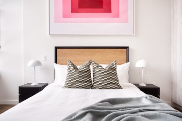 Modern bedroom with bed, wood headboard, contemporary art, black nightstands, white lamps, gray blanket, pillows.